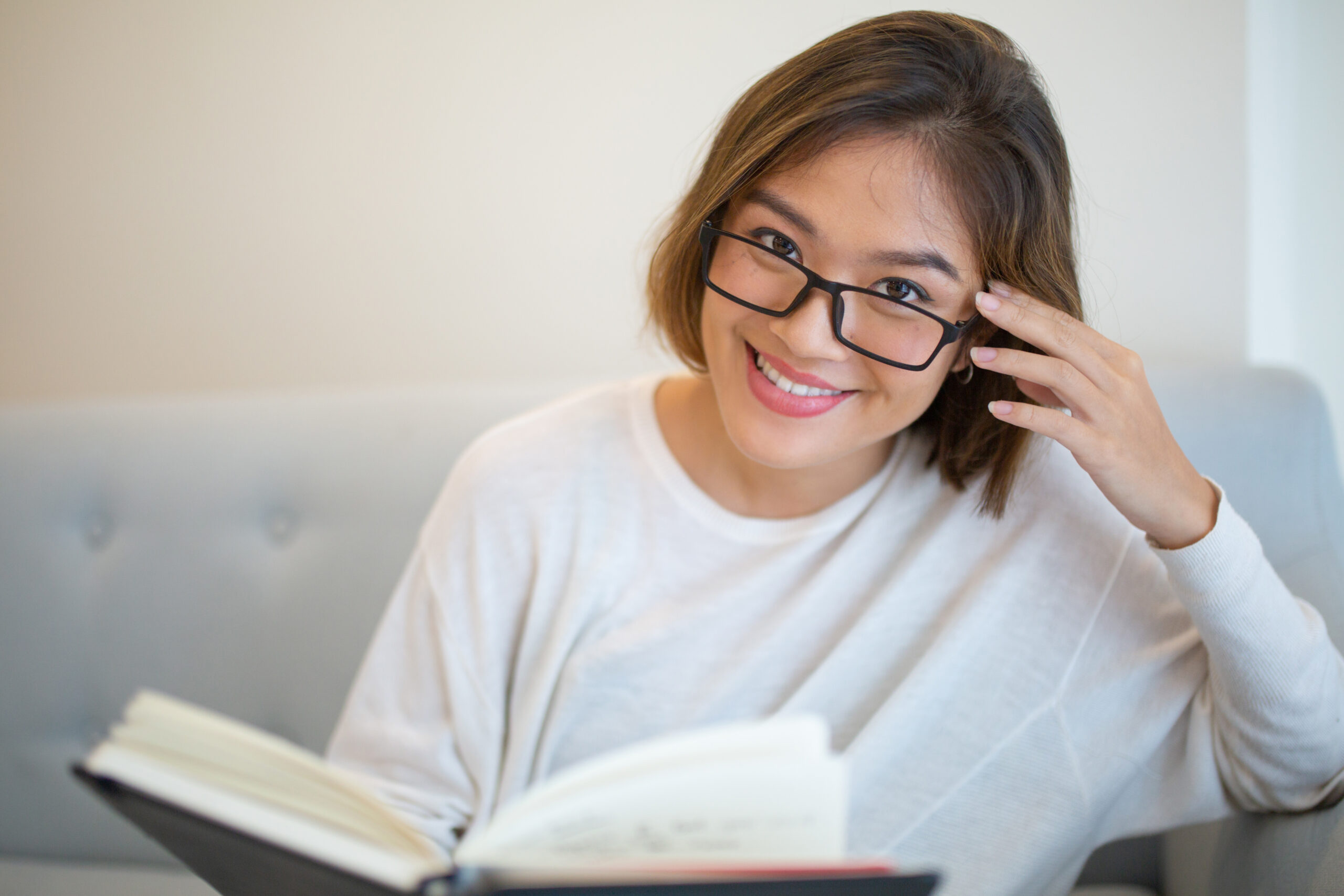 Smiling young woman reading book on couch at home. Pretty Asian lady looking at camera, sitting and holding book. Education or literature concept. Front view.