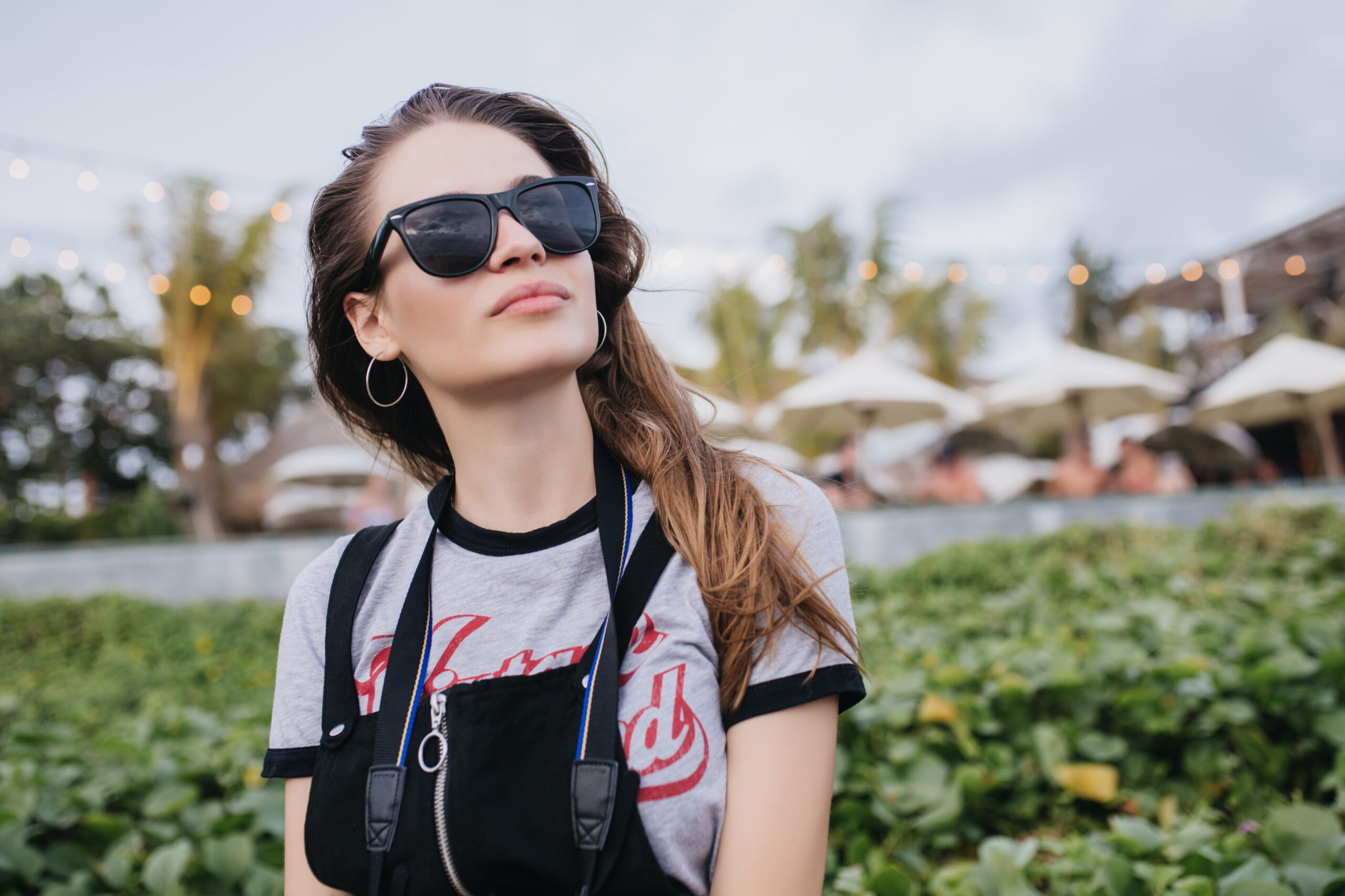 Winsome girl in black sunglasses looking at sky on nature background. Outdoor shot of fashionable brunette woman spending time outdoor in evening.